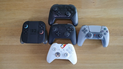 The Switch 2 Pro controller (centre) compared to the Switch 1 Pro controller (top), the PS5 controller (right), the Xbox controller (bottom), and the Joy-Con 2 controller in its charging cradle (left).
